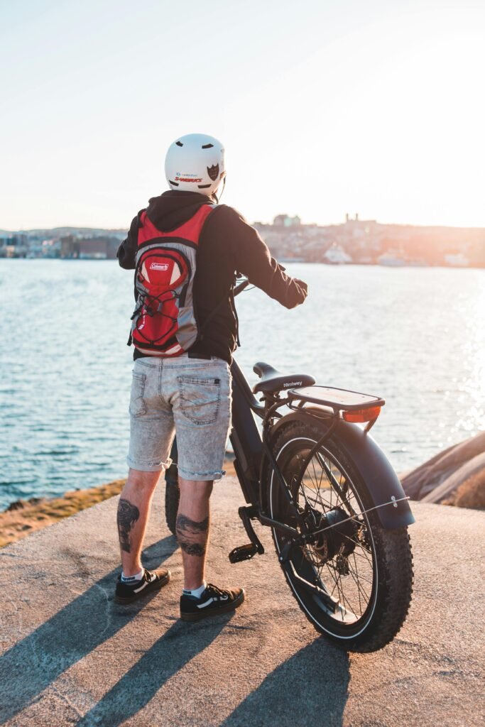 Full body back view of anonymous rider with backpack standing on waterside with bicycle near rippling river against shore with buildings